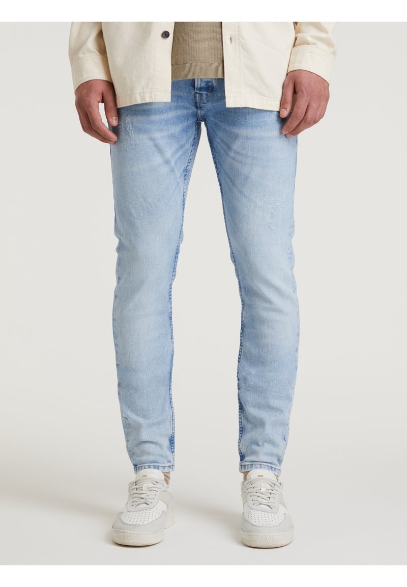 Ljust blå, slim-fit jeans med en urtvättad finish och subtila slitningsdetaljer. Bärs med vita och grå sneakers.