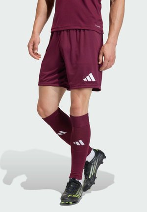 Mężczyzna w bordowych szortach piłkarskich, dopasowanych podkolanówkach z białymi logo i czarnych korkach z neonowozielonymi akcentami.