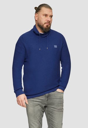 Marineblauwe pullover hoodie met een hoge kraag, koordjes en een klein logopatch, gecombineerd met lichtgrijze denim jeans.