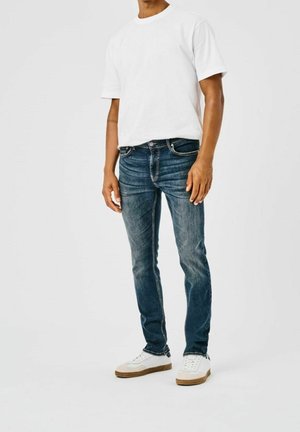 Mężczyzna stojący, ubrany w białą koszulkę z krótkim rękawem, dopasowane niebieskie dżinsy i białe trampki z gumowymi podeszwami na jednolitym jasnym tle.