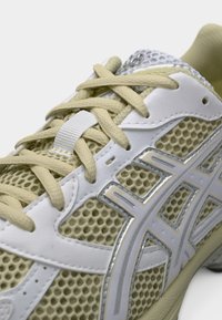 Ανοιχτό μπεζ αθλητικό παπούτσι με πλέγμα στο επάνω μέρος, λευκές λεπτομέρειες και αντανακλαστικά στοιχεία· διαθέτει επίπεδα κορδόνια και υφή στη σόλα.