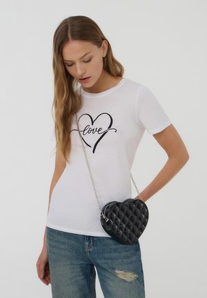 T-shirt bianca in cotone con stampa nera "love" in design a cuore, abbinata a una borsa a tracolla nera a forma di cuore trapuntata con tracolla a catena.