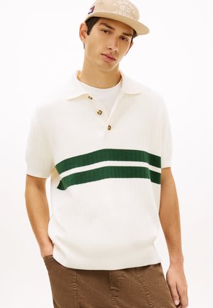 Junger Mann trägt beige Tommy Jeans Mütze, weißes kurzärmeliges Strick-Poloshirt mit grünen Streifen und braune Hose, posiert mit einer Hand in der Tasche.