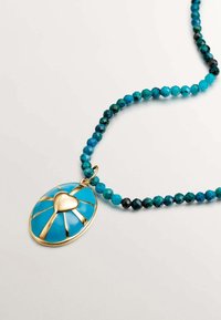 Collar de cuentas turquesas con un colgante ovalado que presenta un diseño de corazón dorado y acentos lineales. Cuentas texturizadas en diferentes tonos de azul.