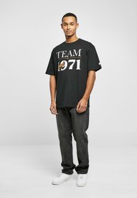 Tricou negru cu grafica "TEAM 1971" în alb și portocaliu, purtat cu blugi negri și adidași albi, stând pe un fundal neutru.