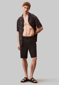 Camicia nera a maniche corte con motivo geometrico bianco, abbinata a pantaloni corti neri e sandali neri. Il modello è in piedi su uno sfondo grigio.