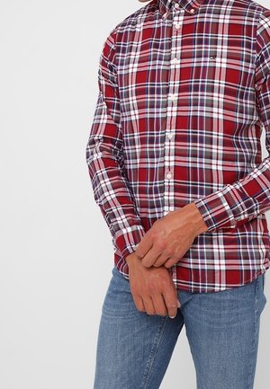 Personne portant une chemise à carreaux rouge, blanche et bleue à manches longues boutonnée, avec un jean bleu, les mains jointes devant.