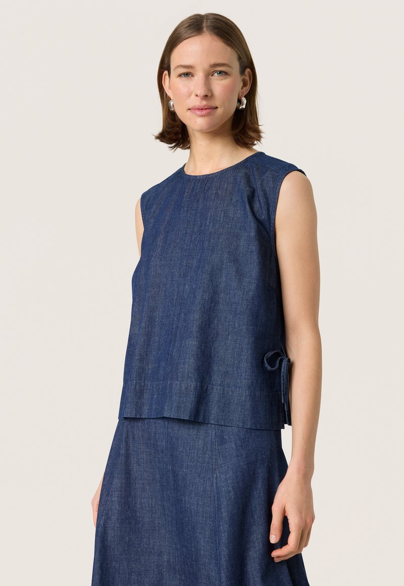 Denim sleeveless top featuring a round neckline, subtle texture, and a side tie detail. Color is deep blue; loose-fitting design.