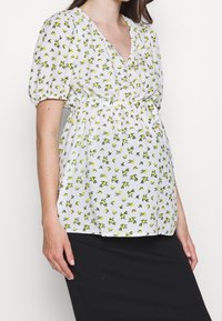 Blouse blanche à motif floral jaune et vert, manches courtes ballon, col en V et taille élastique, associée à une jupe noire.
