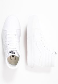 Białe trampki w stylu high-top z teksturowanym wykończeniem, płaskimi sznurówkami i wyściółką przy kołnierzu. Posiadają markowy język i przeszycia na bocznych panelach.