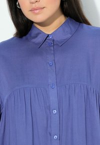 Camicia button-up viola con colletto, caratterizzata da piccoli bottoni coordinati e tessuto arricciato sotto il busto, realizzata in materiale leggero.
