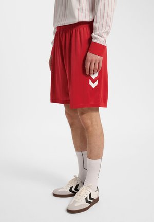 Mężczyzna w czerwonych sportowych spodenkach z białymi prążkami w kształcie litery V, białych skarpetach za kostkę, białych trampkach i białej koszulce z długim rękawem z czerwonymi paskami.
