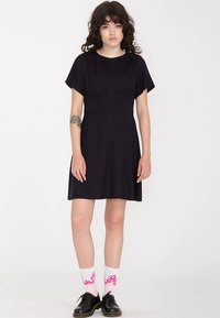 Czarna sukienka z krótkim rękawem, dopasowanym górnym elementem i rozkloszowanym dołem. Gładki materiał, długość do kolan, zestawiona z różowymi skarpetkami w płomienie i czarnymi butami.