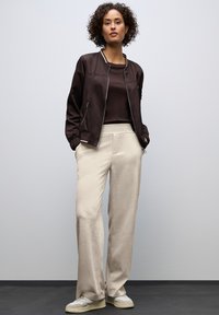 Brązowa kurtka bombowca z żebrowanymi akcentami, zestawiona z beżowymi spodniami o szerokich nogawkach. Model nosi jasne sneakersy. Gładkie tło.