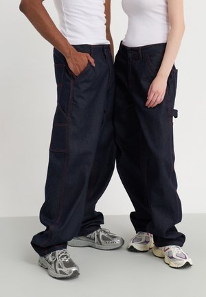 Deux personnes debout côte à côte portant des hauts blancs, des pantalons en jean sombre amples et des baskets de sport sur un fond clair uni.