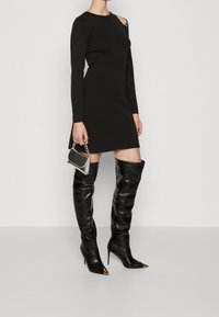 Svart långärmad klänning med asymmetrisk axeldetalj och dekorationer, kombinerad med glänsande svarta stövlar som går över knäna och en liten handväska.