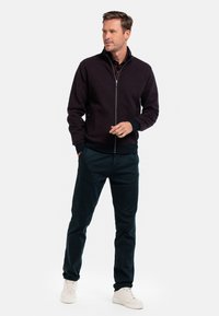 Bordeauxrode ritsjack met een hoge kraag, ribbelmanchetten en een ribbelzoom. Gecombineerd met donkergroene broek en witte sneakers, wat een casual outfit creëert.