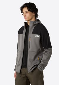 Szara i czarna kurtka z kapturem z zamykanymi kieszeniami, wykonana z miękkiego materiału. Na piersi znajduje się logo The North Face, a suwakowe uchwyty są w kolorze pomarańczowym.