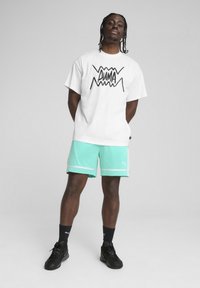 T-shirt blanc avec un logo graphique noir "PUMA" et un short vert avec des accents blancs. Des baskets noires et des chaussettes complètent la tenue.