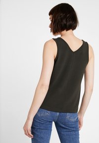 Ærmeløs top i mørkegrøn stof med V-hals, glat tekstur og tætsiddende design. Bagpå er der en syningdetalje. Parret med blå jeans.