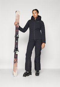 Black insulated jacket with horizontal stitching, paired with matching black ski pants. Ski boots and a ski with a multi-tone design are included.