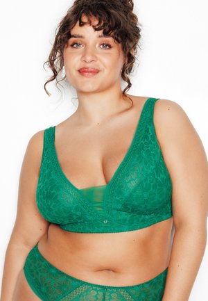 Vrouw met krullend haar, gekleed in een groene bloemenkant beha en bijpassend hooggetailleerd ondergoed tegen een witte achtergrond.