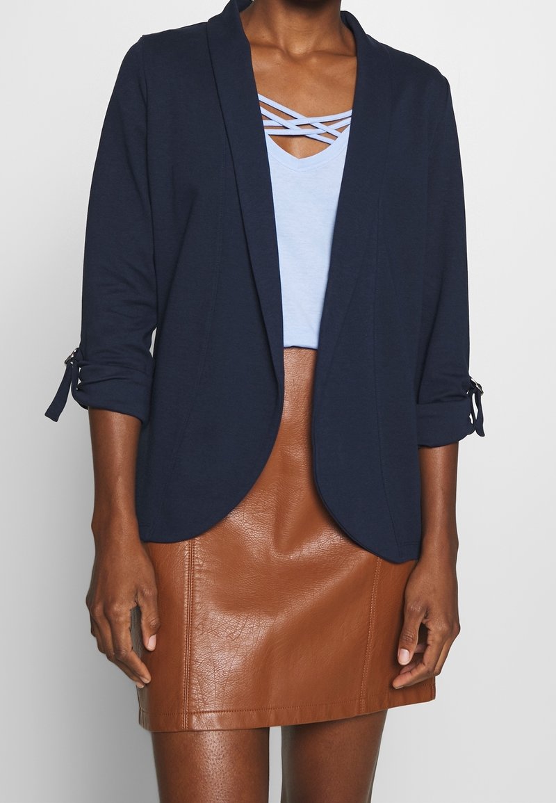 Femme portant un blazer bleu marine avec manches retroussées, un haut bleu clair à bretelles croisées et une jupe en cuir marron, visible des épaules jusqu'à mi-cuisse.