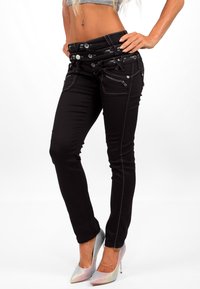 Zwarte skinny jeans met meerdere ritssluitingen, zilveren knopen en contrasterende stiksels, gecombineerd met metallic hoge hakken.