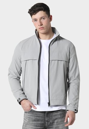 Joven con chaqueta gris claro con cremallera sobre una camiseta blanca y vaqueros desgastados, de pie con una mano en el bolsillo, fondo neutro.