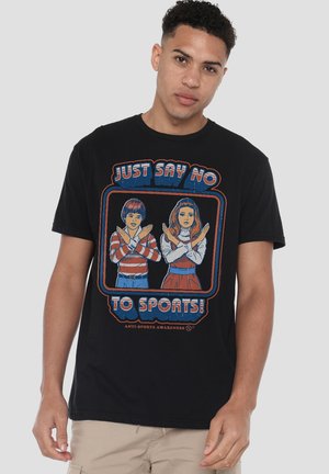 Young man wearing black T-shirt with retro illustration of two children crossing arms and text "Just Say No To Sports!"