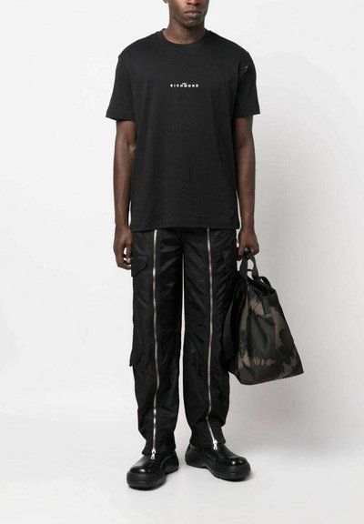 T-shirt nera in cotone con un logo, abbinata a pantaloni cargo neri con cerniere argento e scarpe platform nere. Tiene una borsa a fantasia mimetica.