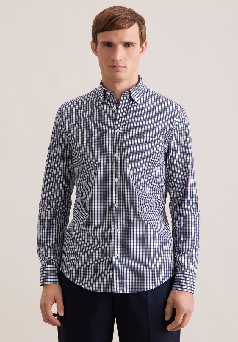 Jeune homme portant une chemise à carreaux bleue et blanche à boutons et un pantalon noir, debout devant un fond beige uni.