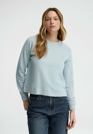 Kvinde med langt blondt hår iført lys blå rundhalset sweatshirt og mørkeblå jeans, stående foran en ensfarvet hvid baggrund.