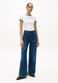 Camiseta blanca de manga corta con ribete azul, que presenta el logo "Tommy Hilfiger", combinada con unos pantalones vaqueros acampanados de tiro alto en azul oscuro. Zapatos negros.