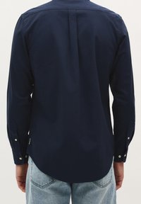 Marineblaues Langarmhemd mit Rückenfalte, Knopfmanschetten und abgerundetem Saum. Der Stoff wirkt glatt mit einem dezenten Glanz.