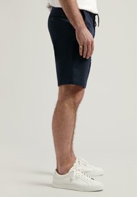 Navy Shorts aus Baumwollmischung, taillierte Passform, seitliche Taschen und ein Kordelzugbund, kombiniert mit weißen Sneakers vor einem neutralen Hintergrund.