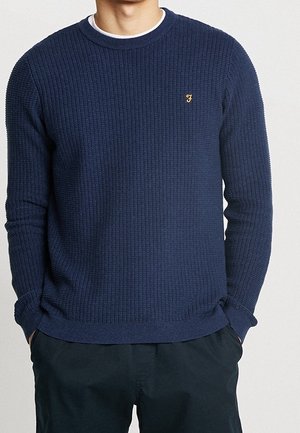 Hombre vistiendo un suéter de punto texturizado azul marino con un pequeño logo dorado "F" y pantalones negros, con las manos en los bolsillos, frente a un fondo liso.