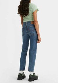 Mujer con cabello largo y ondulado, vestida con una blusa de manga corta verde clara, jeans de cintura alta azul, calcetas blancas y zapatos negros robustos, de espaldas.