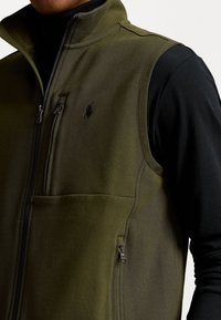 Ciemnozielona kamizelka z polaru z kieszeniami na suwak, wysokim kołnierzem, przeszyciami oraz małym logo na piersi. W zestawie z czarną bluzką.