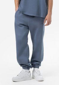 Pantalones de chándal azules hechos de un tejido suave, con puños elásticos, bolsillos laterales y un logo sutil en la pierna. Combinados con zapatillas blancas.