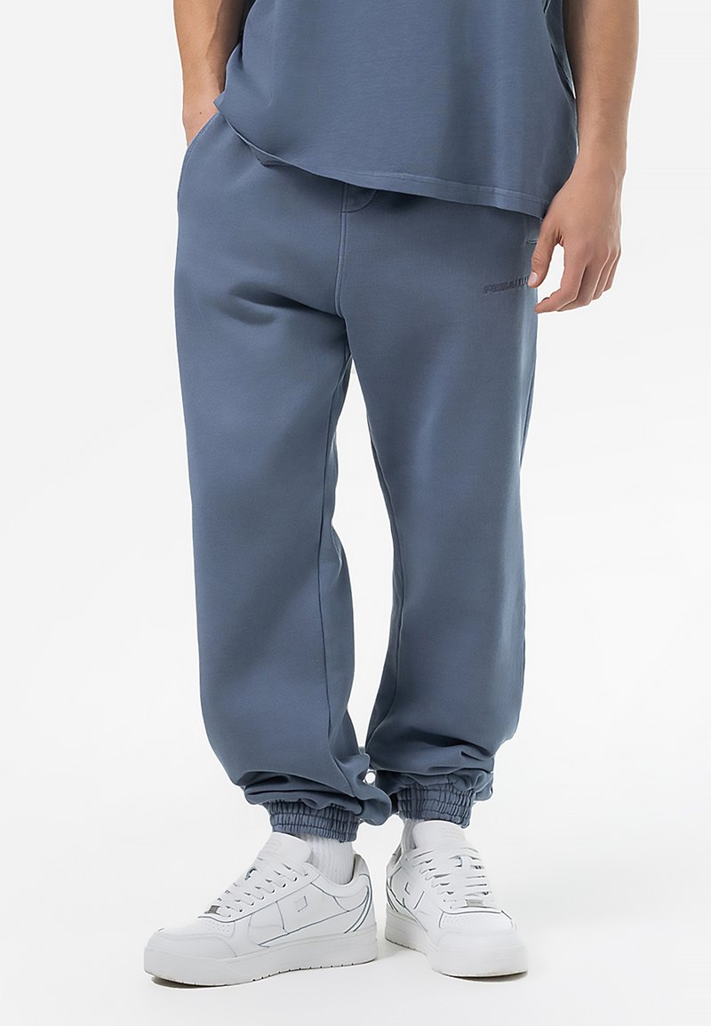 Pantalones de chándal azules hechos de un tejido suave, con puños elásticos, bolsillos laterales y un logo sutil en la pierna. Combinados con zapatillas blancas.