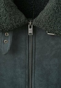 Giacca in faux suede verde scuro con colletto in pelliccia, chiusura con zip e accessori metallici. Presenta cuciture visibili e occhielli.
