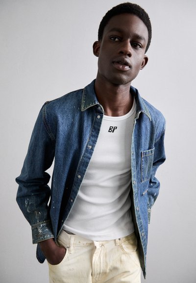 Camisa de denim con manchas desgastadas, cuello abotonado; usada sobre una camiseta sin mangas blanca de canalé; combinada con pantalones claros, ligeramente desgastados.