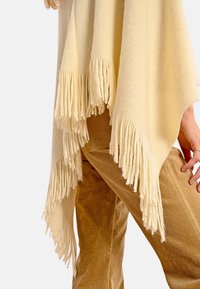 Poncho color crema con bordi frangiati, realizzato in materiale morbido e testurizzato. Indossato sopra pantaloni di velluto a coste color sabbia, che mostrano un design a strati.