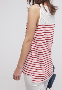 Žena v bezramenném bílém topu s červenými pruhy a krajkovými detaily, oblečená v bílých kalhotách a držící malou tmavě modrou tašku po svém boku.