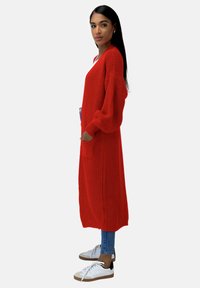 Cárdigan rojo de punto con mangas largas y bolsillos laterales, que presenta un corte holgado y un acabado texturizado, combinado con jeans y zapatillas.