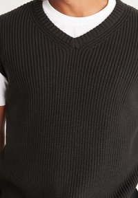 Gilet grigio scuro lavorato a maglia con design a V. Presenta un tessuto testurizzato a coste e una forma aderente. Indossato sopra una maglietta bianca.