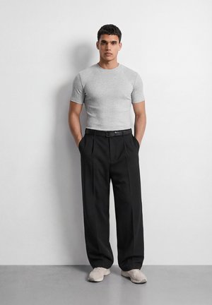 Άνδρας στέκεται μπροστά σε λευκό τοίχο, φορώντας εφαρμοστό, ανοιχτό γκρι κοντομάνικο πουκάμισο, φαρδιά μαύρα παντελόνια με ζώνη και ανοιχτόχρωμα αθλητικά παπούτσια.