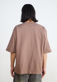 Person med axellångt svart hår som bär en lös kortärmad brun skjorta och blekta mörka jeans, vänd bort på en enfärgad bakgrund.