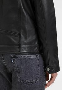 Giacca in pelle nera con una texture liscia, design strutturato e dettagli con bottoni sui polsini, abbinata a jeans in denim scuro.